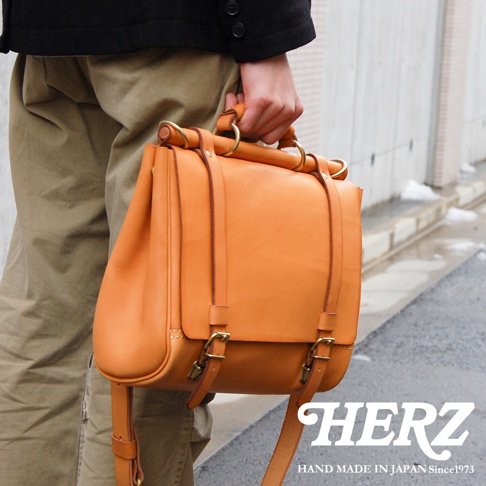 一手提著橘色皮革側背包，包包上有金屬扣環及揹帶，背景為模糊的街道，下方有HERZ品牌標誌和HAND MADE IN JAPAN Since1973字樣。