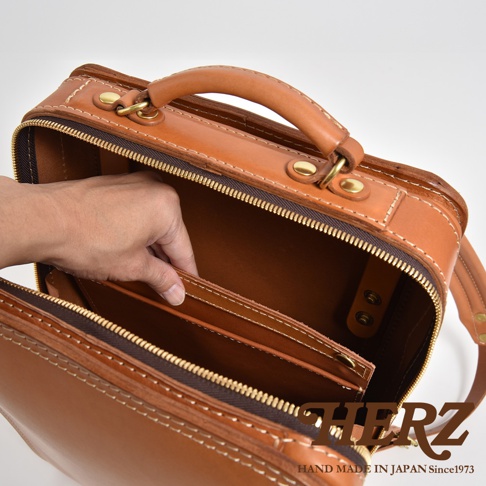 職人手作皮革商務電腦後背包 R-25，採用鞣製皮革材質，呈現自然棕色。包款設有頂部提把、拉鍊開合，內部有隔層設計，可見手部正在放入物品。