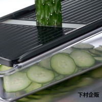 四合一刨絲器，黑色塑膠外框，搭配不鏽鋼刀片，正上方置入一截綠色小黃瓜，下方透明集料盒內有數片均勻切好的小黃瓜片。