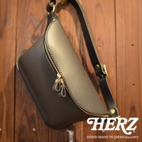 職人手作黑色皮革立體斜背包，設有雙拉鍊開口及皮革拉環，搭配可調式肩帶，掛於木紋牆面。