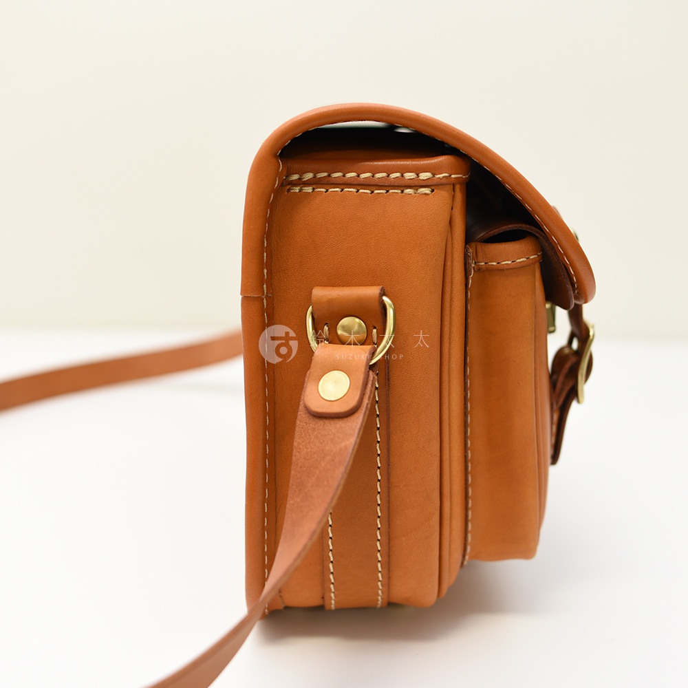 日本HERZ職人手作皮革經典雙扣郵差包，從側面看，咖色皮革包款線條硬挺，配有雙邊金色圓形扣件和皮製背帶。