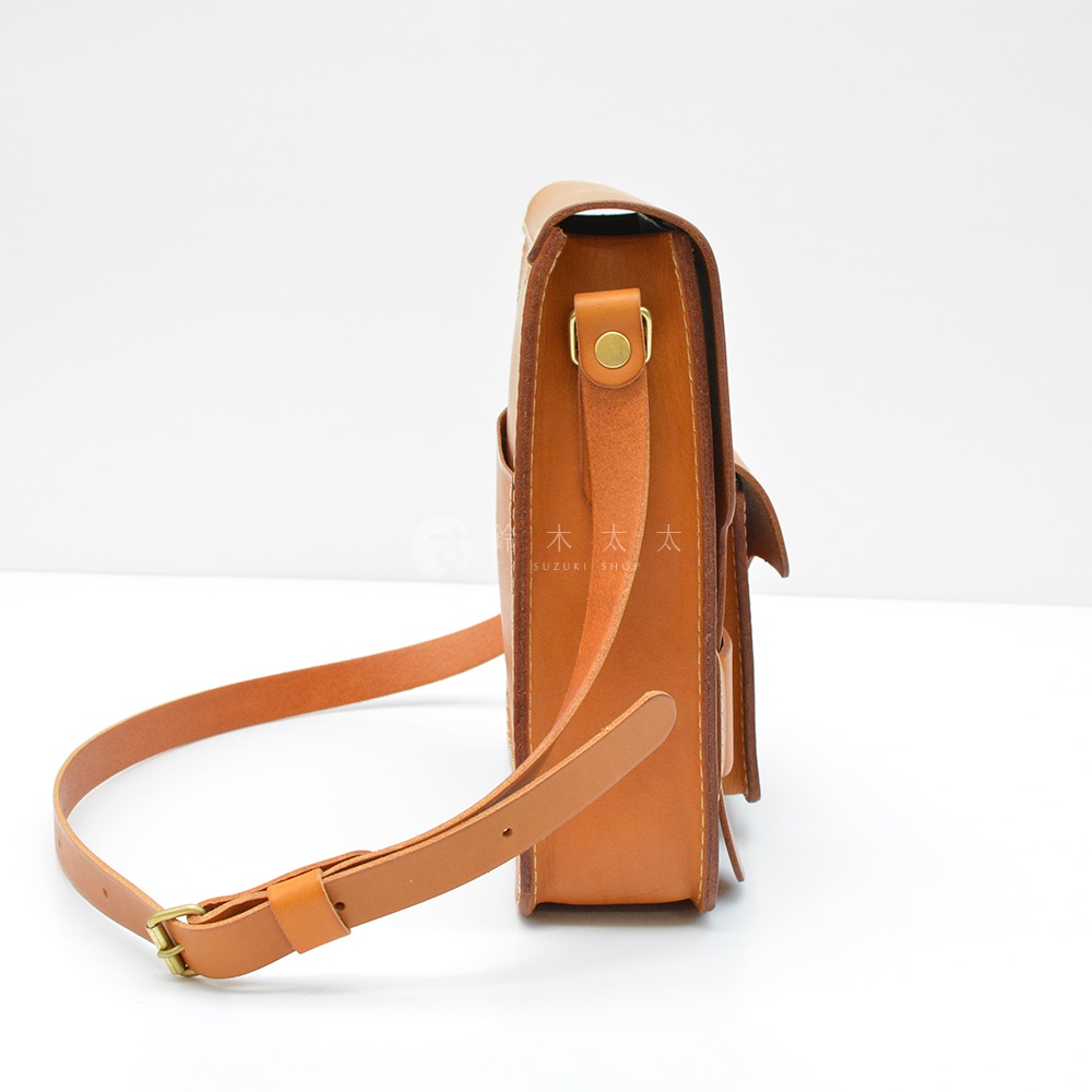 這是一款棕色皮革製成的單肩包，從側邊展示，包身厚實，配有可調節的皮革肩帶和金屬釦環。