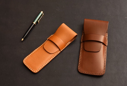 兩款職人手作皮革鋼筆專用收納筆套，一款為淺棕色，另一款為深棕色，皆採用真皮材質，配有縫線及皮革固定帶。一旁擺放一支綠色條紋鋼筆。