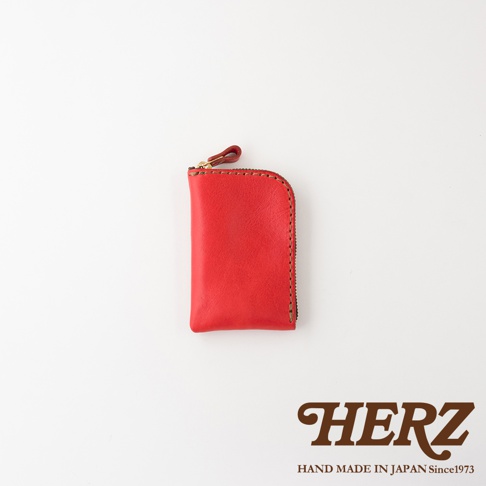 這是一款由日本HERZ職人手作的紅色皮革零錢包，外觀呈現簡約圓角長方形設計，側面配有金色拉鍊與皮革拉環，表面可見細緻的車縫線工藝，整體質感溫潤且厚實，右下角印有HERZ品牌標誌與Hand Made in Japan字樣。