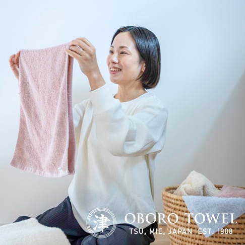 一位女士展示著一條淡粉色的有機棉毛巾，毛巾質地柔軟，可見細緻的織紋。背景有編織籃，內有其他顏色的毛巾。