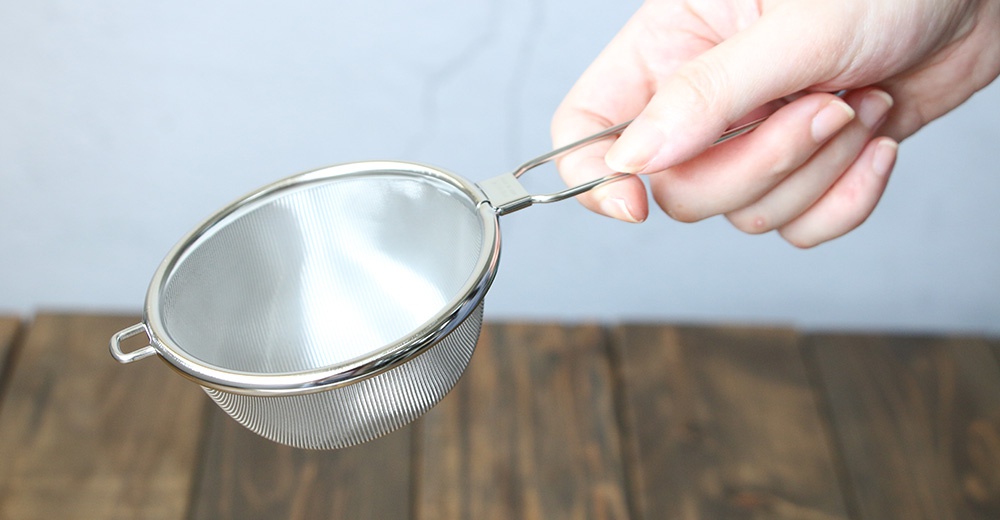 一只手正拿著一個不鏽鋼材質的超細目濾茶器，濾網為銀色，帶有一個銀色的金屬手柄。