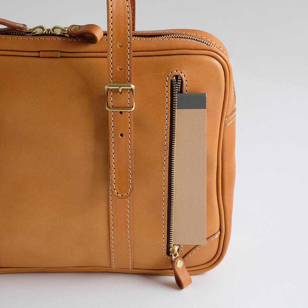 職人手作皮革肩背手提兩用公事包，採用天然淺棕色皮革，搭配金色拉鍊與金屬扣環，車縫線清晰，側邊拉鍊口袋內夾有品牌標籤。