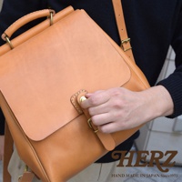 職人手作皮革屋瓦風後背包，採用天然皮革材質，呈現溫潤的淺棕色。方形包款設計，配有可調整的肩帶、頂部提把及金屬旋鈕開關，展現簡約復古風格。