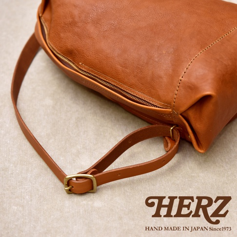 日本職人品牌 HERZ 旗下的 Organ 後背包，選用焦糖棕色義大利 Eufrate 皮革製作，呈現質地柔軟且具自然紋理的皮革質感。影像以斜上方視角捕捉包身側邊的拉鍊開口與金屬扣環背帶細節，呈現簡約且精緻的手作工藝風格，下方印有 HERZ 品牌標誌與成立年份說明。