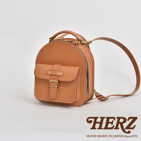 駝色皮革迷你後背包，圓潤造型，正面有翻蓋口袋，以皮帶和金屬扣固定。頂部有提把，側面有拉鍊，附有皮革肩帶。