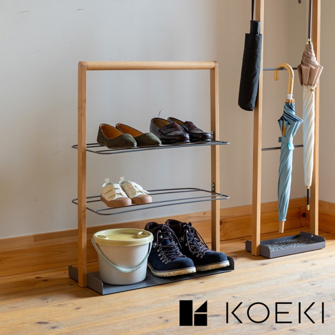 木質框架兩層鞋架，展示著綠色平底鞋、棕色樂福鞋、白色帆布鞋、藍色登山靴，以及白色附黃色蓋子的水桶。旁邊是吊掛著黑色、藍色、米白色和棕色雨傘的衣架。