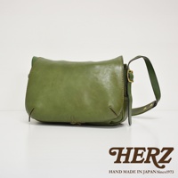 一件復古翻蓋郵差包，採用綠色皮革製成，搭配金屬扣環和可調式背帶。包身線條簡潔，手工縫線細緻，展現皮革的自然質感。