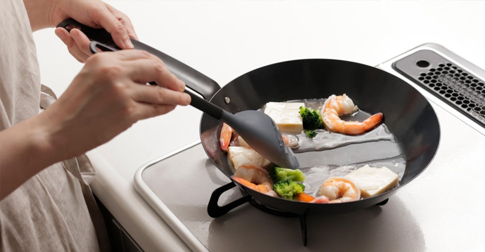 一人正在用灰色耐熱矽膠料理匙在黑色平底鍋中烹調蝦仁、豆腐和花椰菜。