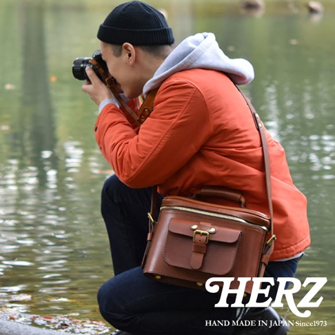 身穿橘色外套、頭戴黑色毛帽的男子蹲在水邊，他正透過鏡頭拍攝眼前的景物，肩上斜背著一個精緻的棕色皮革相機包，包包的右下方有HERZ的白色字樣，下方小字為HAND MADE IN JAPAN Since1973。