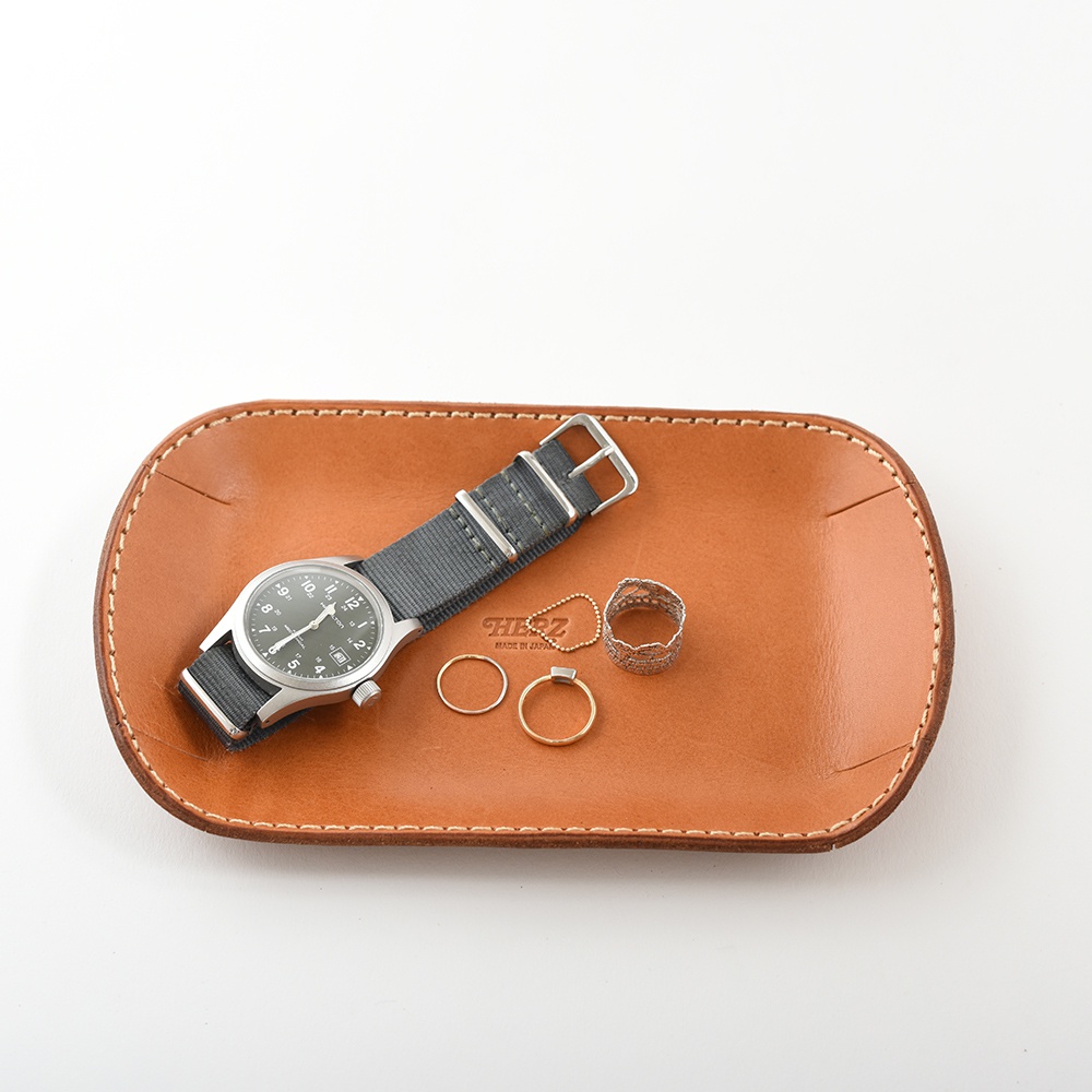 職人手作皮革小物收納盤，淺棕色皮革，搭配灰色錶帶的銀色錶面手錶、兩只金色戒指、一只銀色戒指和一個小項鍊，收納盤邊緣有白色縫線。