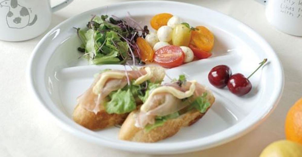 白色琺瑯分隔餐盤，盛有小番茄、莫札瑞拉起司球、沙拉，以及兩份火腿生菜小點心，盤邊有兩顆櫻桃。