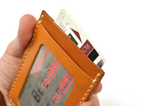 職人手作皮革直式票券收納夾，呈現天然皮革的焦糖色澤，側邊以米白色線縫合。夾內放置了多張票券或卡片，其中可見一張印有「RO」字樣的卡片。