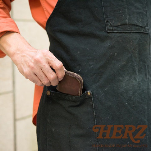 身穿黑色工作圍裙的人將一個深咖啡色的圓弧形手工皮革零錢包放入圍裙口袋中，影像右下角印有日本品牌HERZ的標誌。