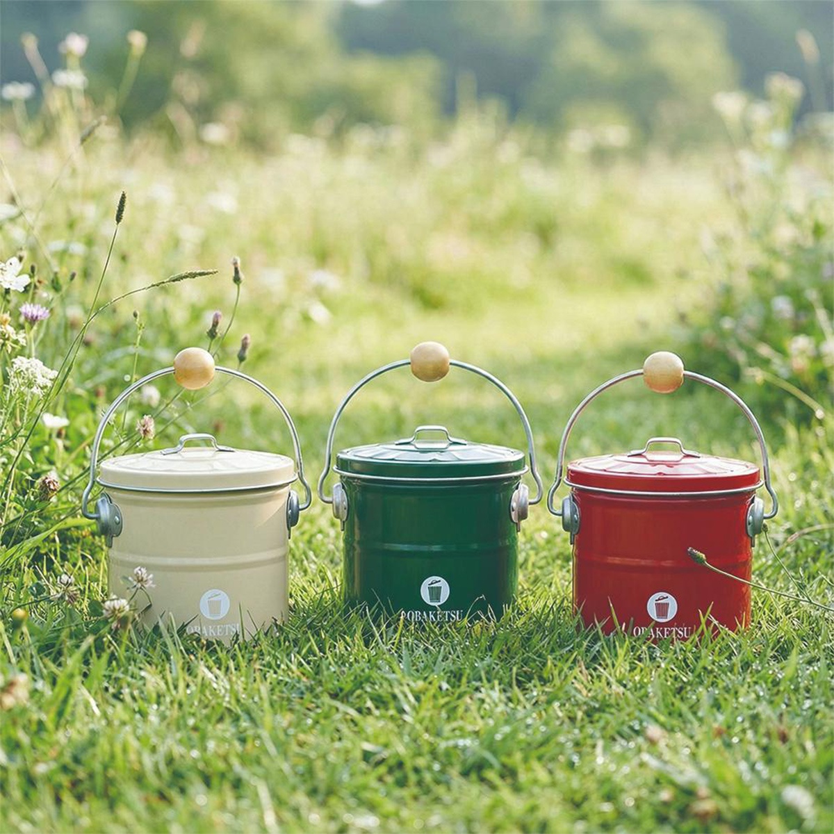 綠色、紅色和米黃色的OBAKETSU手工製蚊香桶，放置於草地上，每個桶子都有金屬提把和木製把手，上面印有品牌Logo。