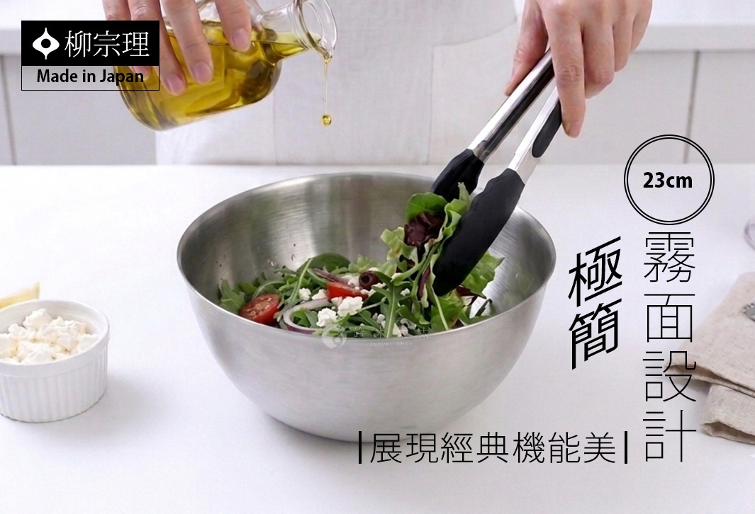 柳宗理品牌的不鏽鋼霧面調理盆，搭配金屬柄矽膠頭食物夾。畫面中央為一個直徑23公分的圓形不鏽鋼盆，正在盛裝生菜沙拉，一雙手持食物夾拌入橄欖油，左側有一小碗起司，背景為極簡白桌與廚房環境，展現經典功能美學。