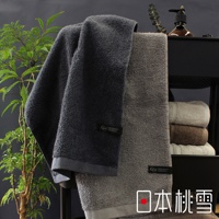 兩條厚實的男人大毛巾，一條為深灰色，另一條為淺灰色。毛巾質地蓬鬆，皆有品牌標籤，背景有綠植與其他顏色毛巾堆疊。