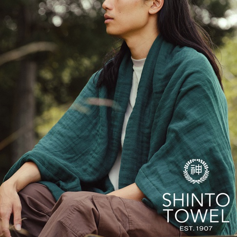 身著一件深綠色2.5層紗材質的隨身暖毯，款式寬鬆，觸感輕柔，搭配白色內衣與棕色長褲。