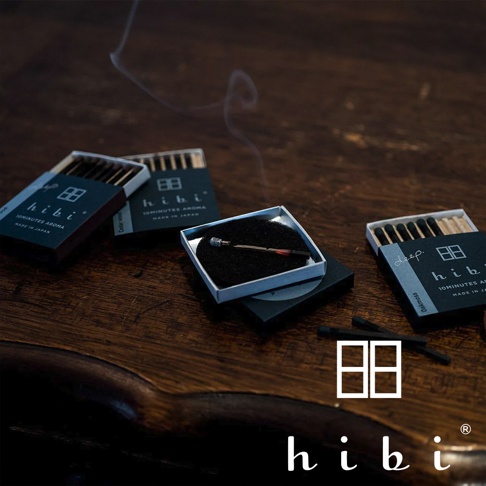 木質桌面上擺放著三盒 hibi 香氛火柴禮盒，其中一根火柴正在專用黑色燃燒墊上燃燒並散發細微煙霧。圖片展現了極簡風格的包裝設計，整體呈現溫潤且精緻的質感，右下角標註有 hibi 品牌標誌。