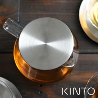 Carat茶壺600ml，透明玻璃壺身裝有琥珀色液體，搭配霧面不鏽鋼圓形壺蓋，壺嘴為玻璃材質，放置於深色木紋桌面上。