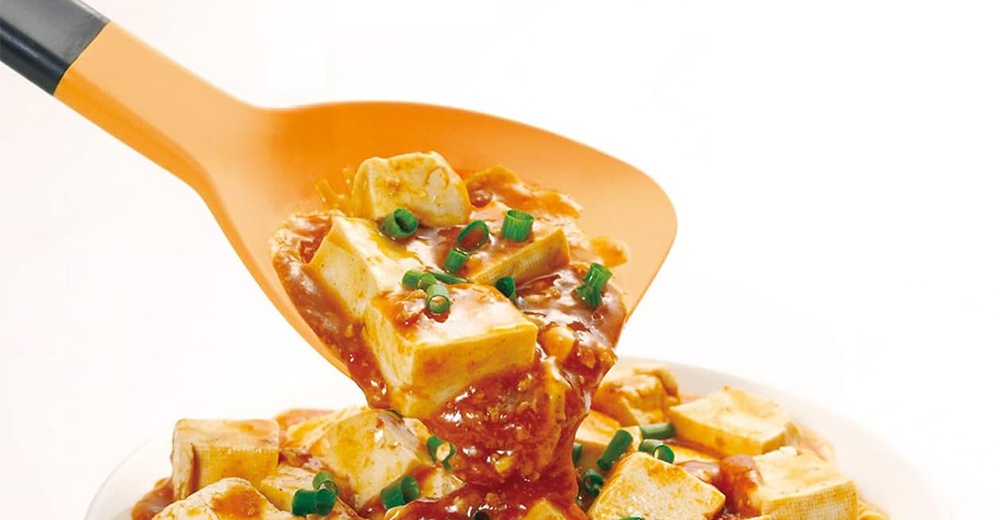 以橘色鍋鏟盛起一勺紅燒豆腐，豆腐塊浸潤在醬汁中，點綴著蔥花。