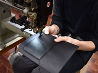 職人正在使用工業縫紉機縫製黑色皮革翻蓋迷你單肩包。包袋採用光滑皮革製成，邊緣有精細的縫線。