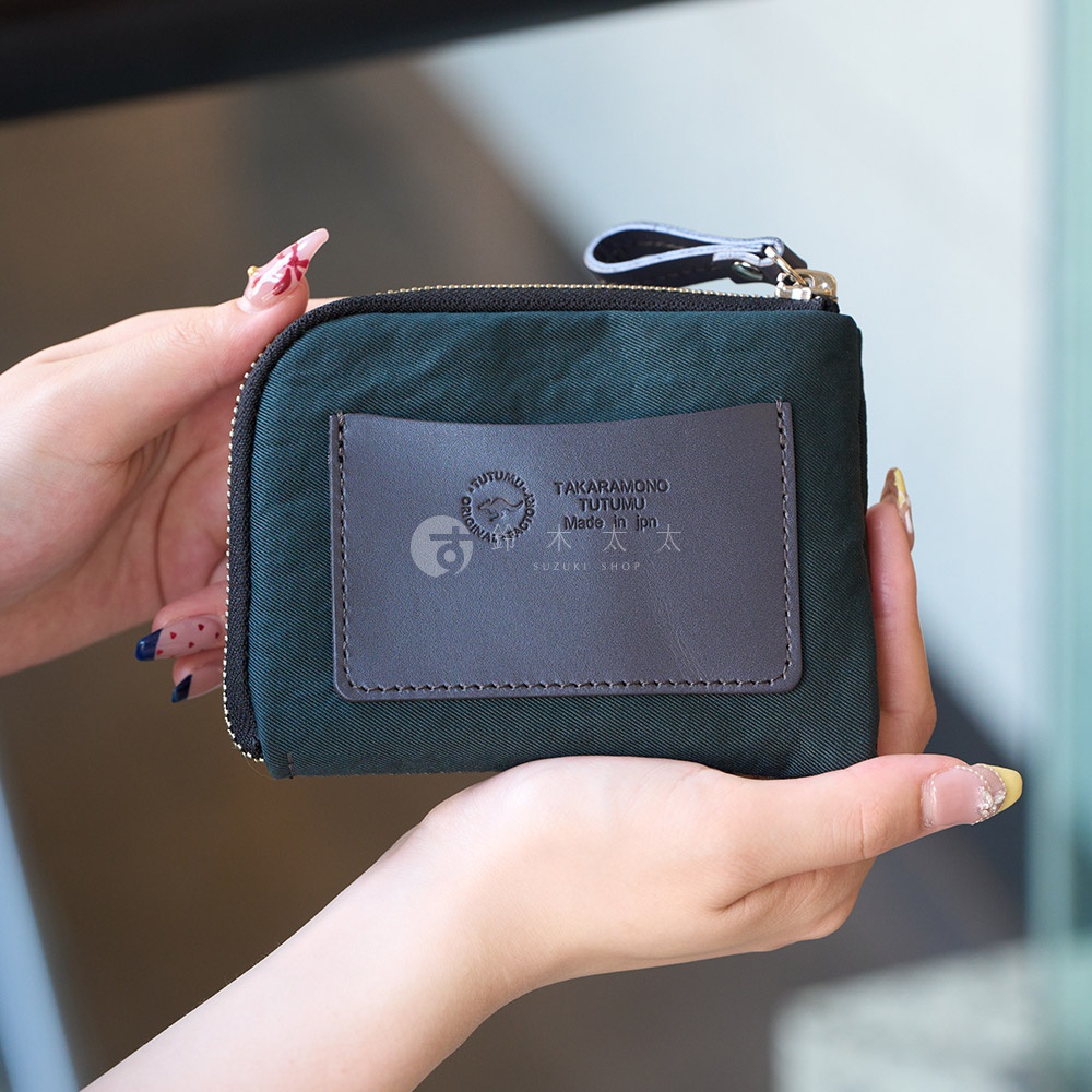 一個深綠色的迷你錢包，上面有一個咖啡色的皮革標籤，上面印有「TAKARA MONO TUTUMU Made in Jpn SUZUKI SHOP」的字樣，由一雙手拿著。