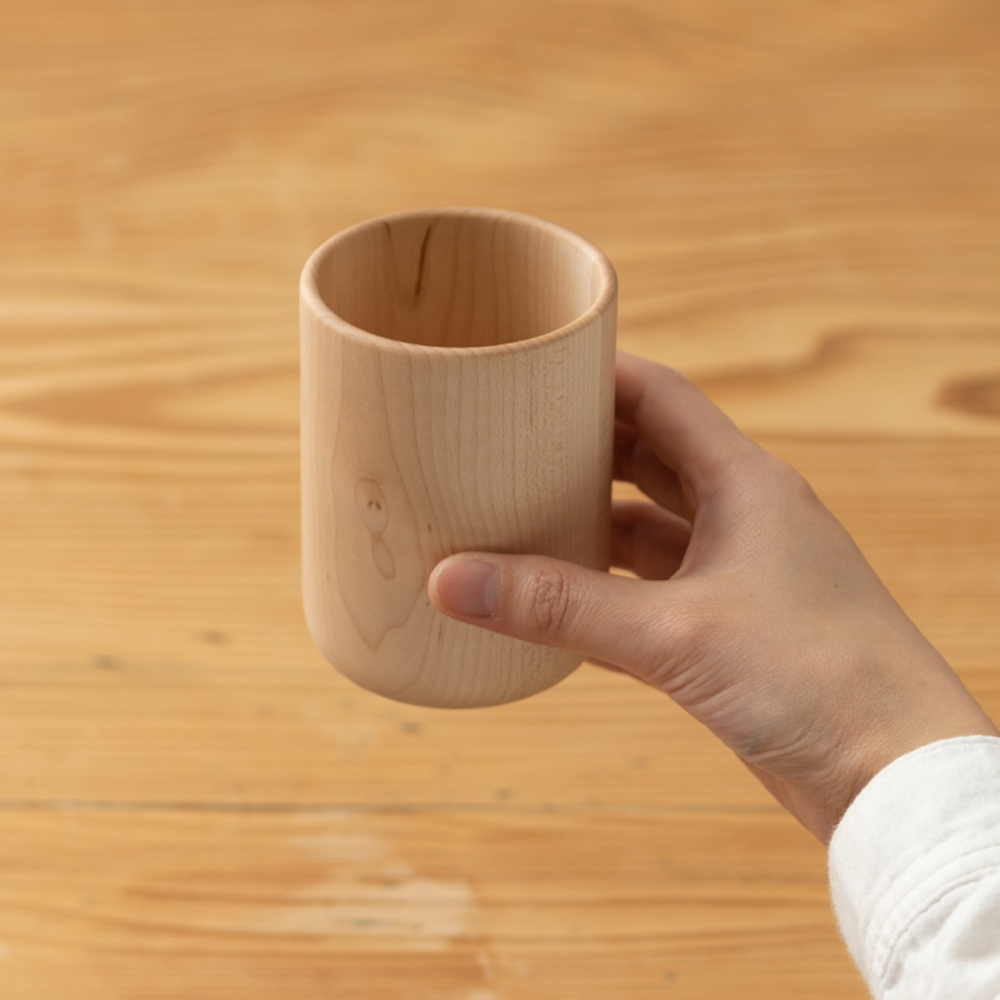 一只手拿著一個淺木色的楓木水杯，杯子呈現圓柱形，邊緣光滑，內壁清晰可見。背景為木紋桌面。