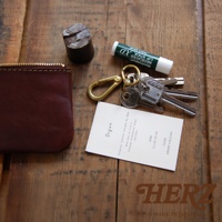 酒紅色職人手作皮革Organ拉鍊式小物收納袋，搭配金色金屬拉鍊。旁邊有鑰匙圈、幾串鑰匙、一條護唇膏，以及一張印有品牌資訊的白色小卡。