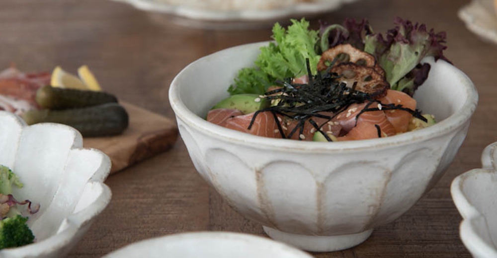 一個裝有鮭魚、酪梨、海帶和生菜的白色陶瓷丼碗，碗身帶有凹凸紋理，碗旁有醃黃瓜和檸檬。