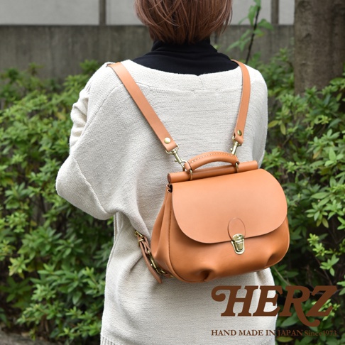 一位女性背著日本HERZ職人手作皮革後背包，這款屋瓦風迷你背包採用淺棕色皮革製作，搭配黃銅金屬扣環與可拆卸調整背帶，外型呈現簡約且溫潤的質感，下方印有HERZ品牌標誌。