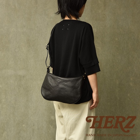 一位模特身著黑色上衣和米色褲子，斜挎一個黑色半月形單肩包。包袋由皮革製成，表面呈現自然的紋理，有金屬搭扣和肩帶調節扣。