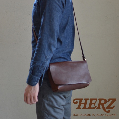 一個身穿藍色牛仔襯衫的人側身站立，斜挎著一個深棕色皮革信使包，包的蓋子向上翻起，露出包的內部，包的右下角有HERZ的標誌和HAND MADE IN JAPAN Since1973的文字。