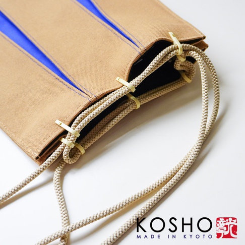 京都KOSHO品牌ougi帆布信玄袋的特寫視角，展示駝色帆布材質配搭鮮明藍色內襯的設計，袋口處綴有米色編織繩與特殊的象牙白塑料扣件細節，整體呈現簡約日式風格。