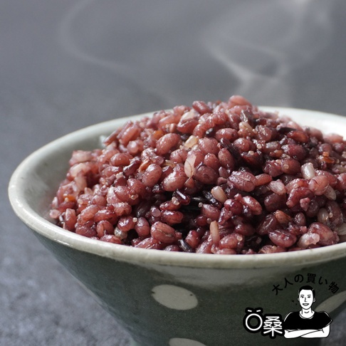 一碗裝滿深紅色十六穀米的陶瓷碗，米飯表面冒著輕煙，碗身有綠色和白色圓點圖案。