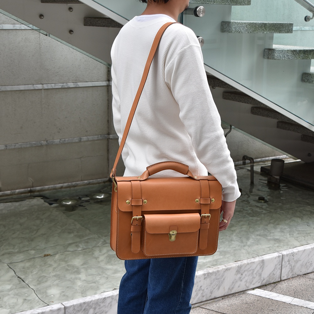 職人手作皮革學院風兩用商務公事包，呈現自然棕色，以高品質皮革製成，設計簡潔大方。包款配有頂部提把、可調節肩帶，以及前袋和金屬扣環，兼具實用與風格。