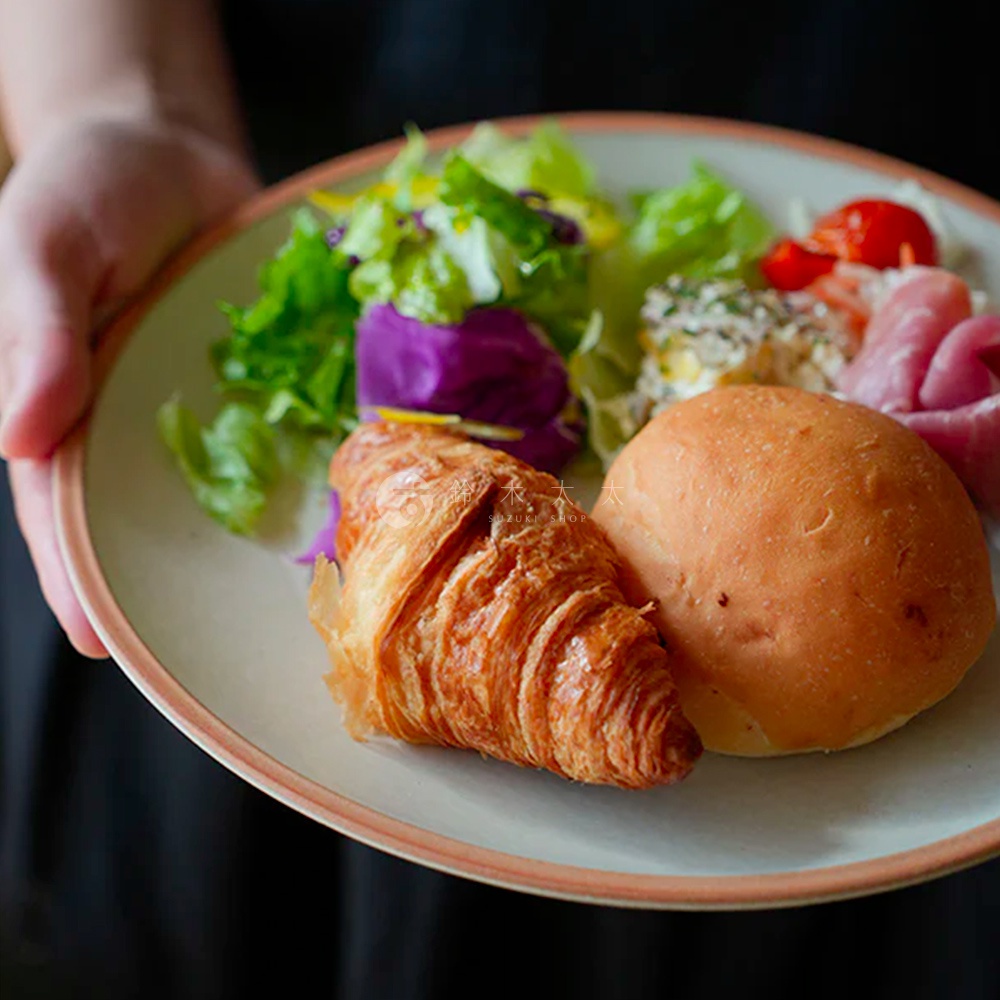 PURE 素燒百搭陶瓷主菜盤，盤身為淺米色，邊緣帶有細緻的粉色滾邊，盤中盛有可頌、小餐包、沙拉、番茄與火腿片。