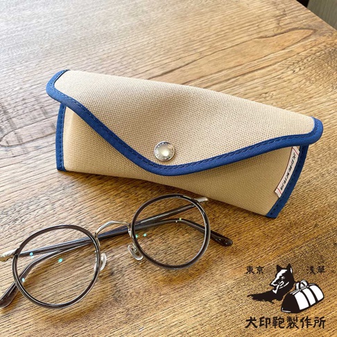 犬印鞄製作所的圓框眼鏡與淺卡其色帆布眼鏡盒，眼鏡盒飾有藍色滾邊及金屬鈕扣，盒內印有日文文字。