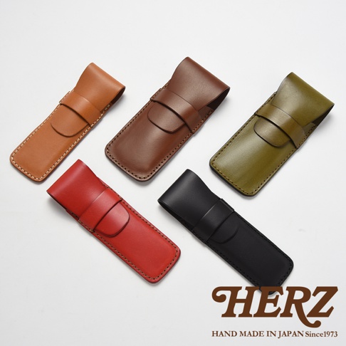 這是一張高視角拍攝的商品圖，展示了日本HERZ職人手作的皮革鋼筆收納筆袋。畫面中共有五款不同顏色的皮革筆袋，分別為焦糖色、棕色、橄欖綠、紅色與黑色。這些筆袋設計簡約，採用經典的皮革扣帶固定結構，縫線工整細緻，展現出高質感的皮革工藝。右下角印有HERZ品牌標誌及相關文字。