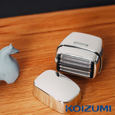 放在木質桌面上的銀色金屬質感KOIZUMI五刃勁剃刀，右側為主體並露出銀色金屬刀網，左前方放置著拆下的保護蓋，旁邊有一個灰色的裝飾物，整體造型簡約時尚。