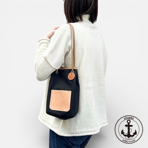 一名女子從背後展示一個黑色束口帆布包，包包上有一個淺棕色皮革前袋和一個帶有錨圖案的皮革吊飾，她身穿白色高領毛衣。