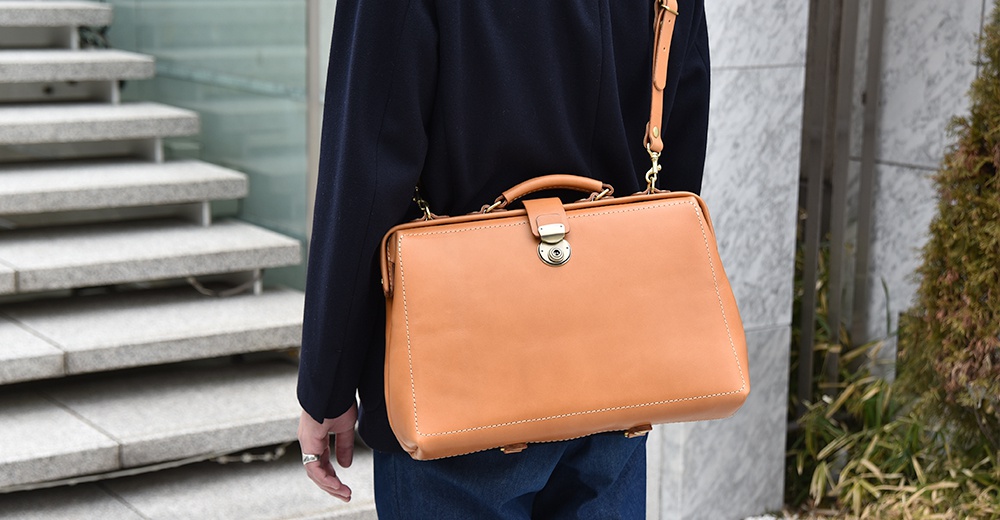 淺棕色皮革製成的手提包，具備可拆卸的肩背帶，包面有縫線裝飾，正面中央有金屬扣環。