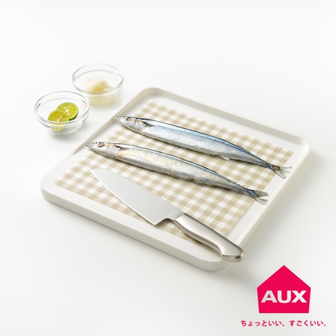 白色防溢大砧板上，鋪著一張米白色格紋的防汙紙，紙上擺放著兩條銀色魚身、魚頭朝左的秋刀魚，以及一把銀色金屬刀。砧板旁有兩個玻璃小碗，分別裝著切片檸檬和白色碎末。