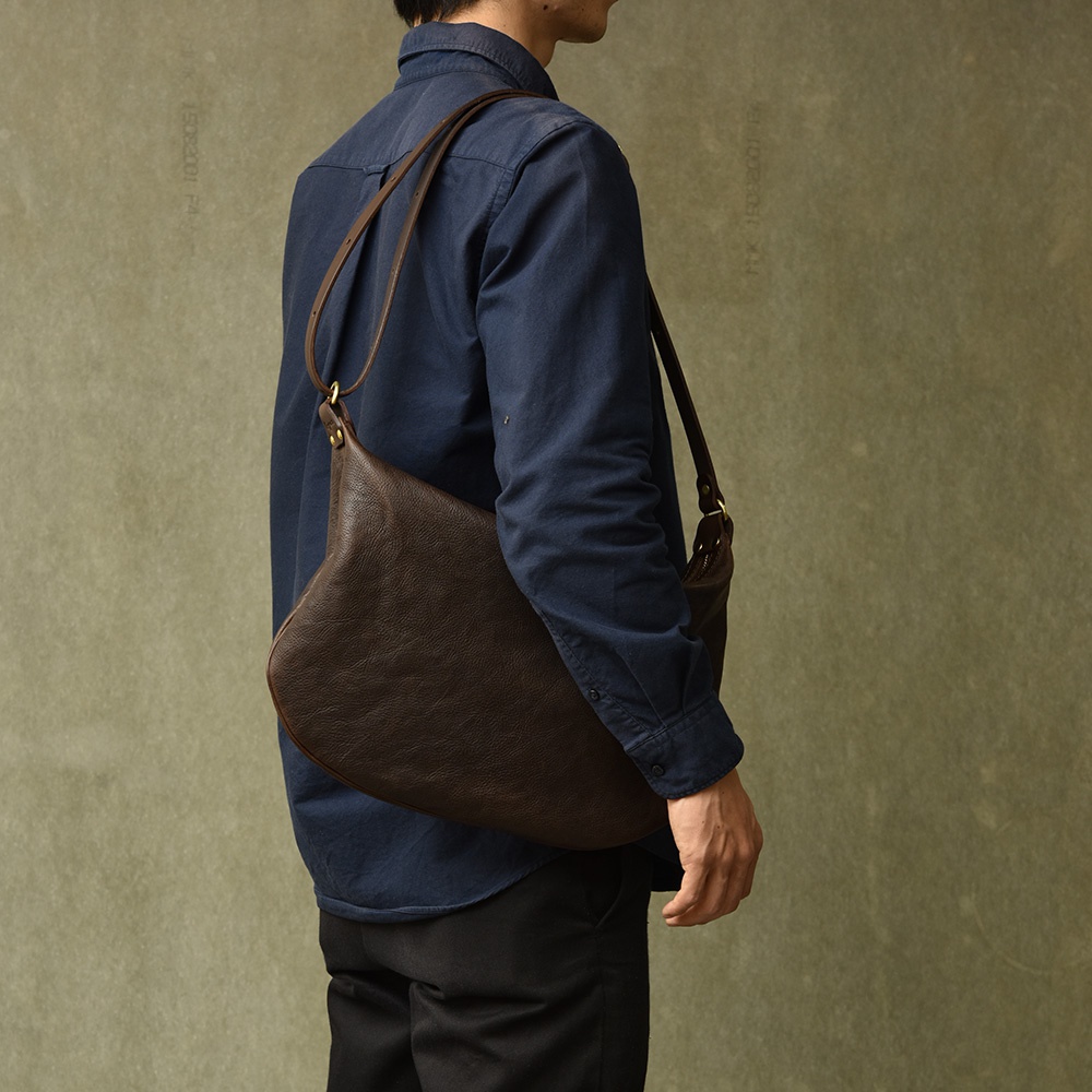 深棕色職人手作皮革Organ單肩半月包，側面展示，包款飽滿，搭配深藍色長袖襯衫與黑色長褲。