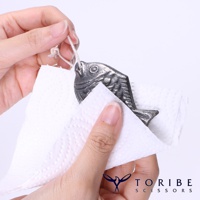 一隻銀灰色金屬製成、帶有麻繩的鯛魚吊飾，正被雙手與白色紙巾擦拭。