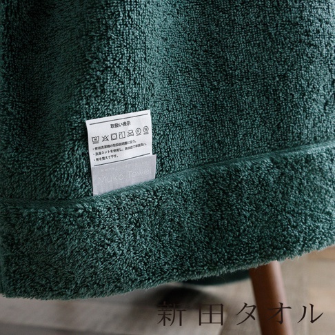 一條深綠色的毛巾織品，局部特寫展示了織物柔軟蓬鬆的毛圈質感。畫面中央縫有一枚白色標籤，上面印有「Muko Towel」品牌名稱及日文洗滌說明符號與保養指引。底部角落處標註有新田塔奧爾的日文字樣。